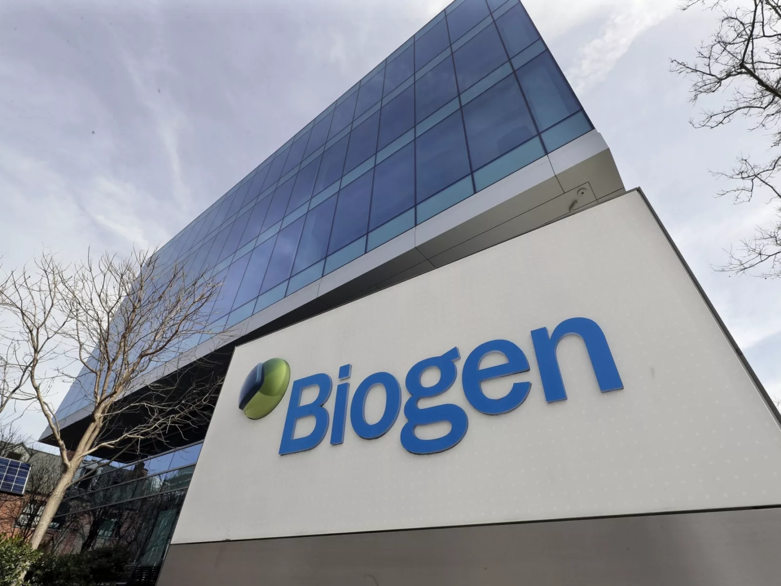 A modern glass building with a prominent Biogen sign in blue, surrounded by bare trees and solar panels, under a cloudy sky.