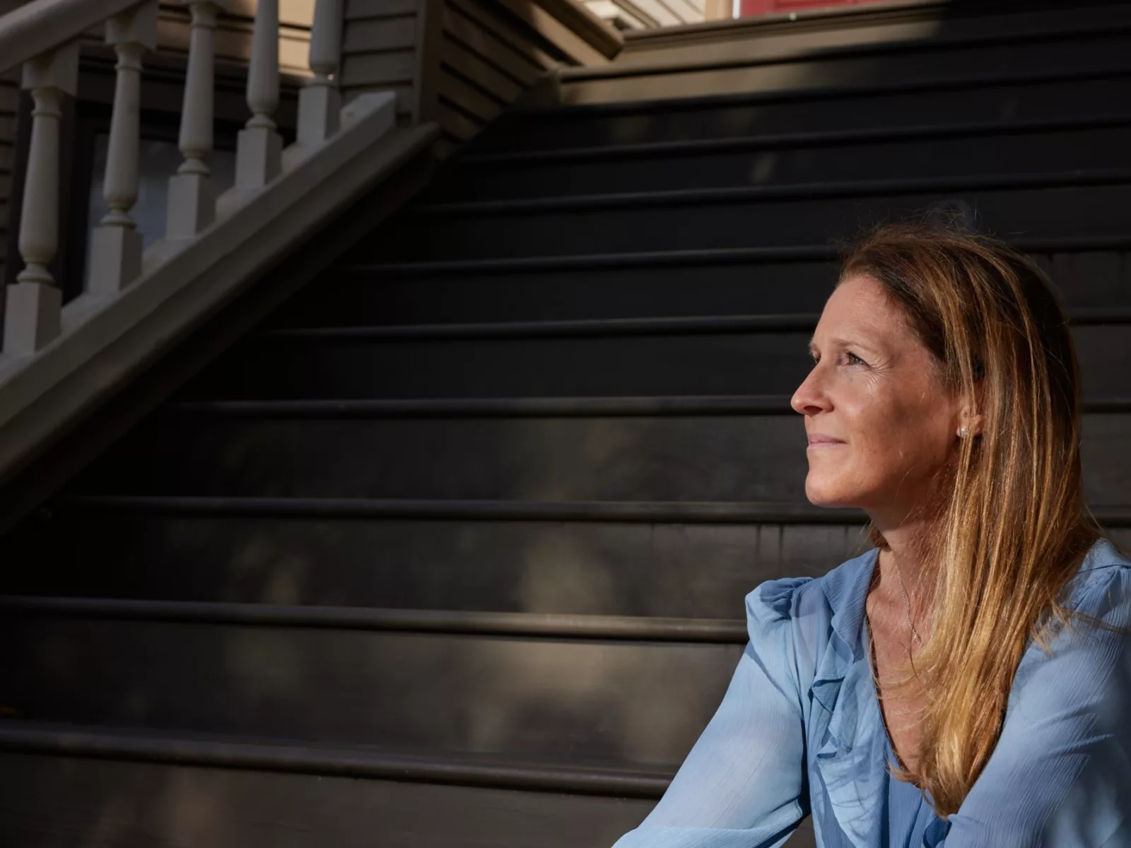 A woman with long, brown hair sits on black wooden stairs, gazing thoughtfully into the distance, dressed in a light blue blouse.
