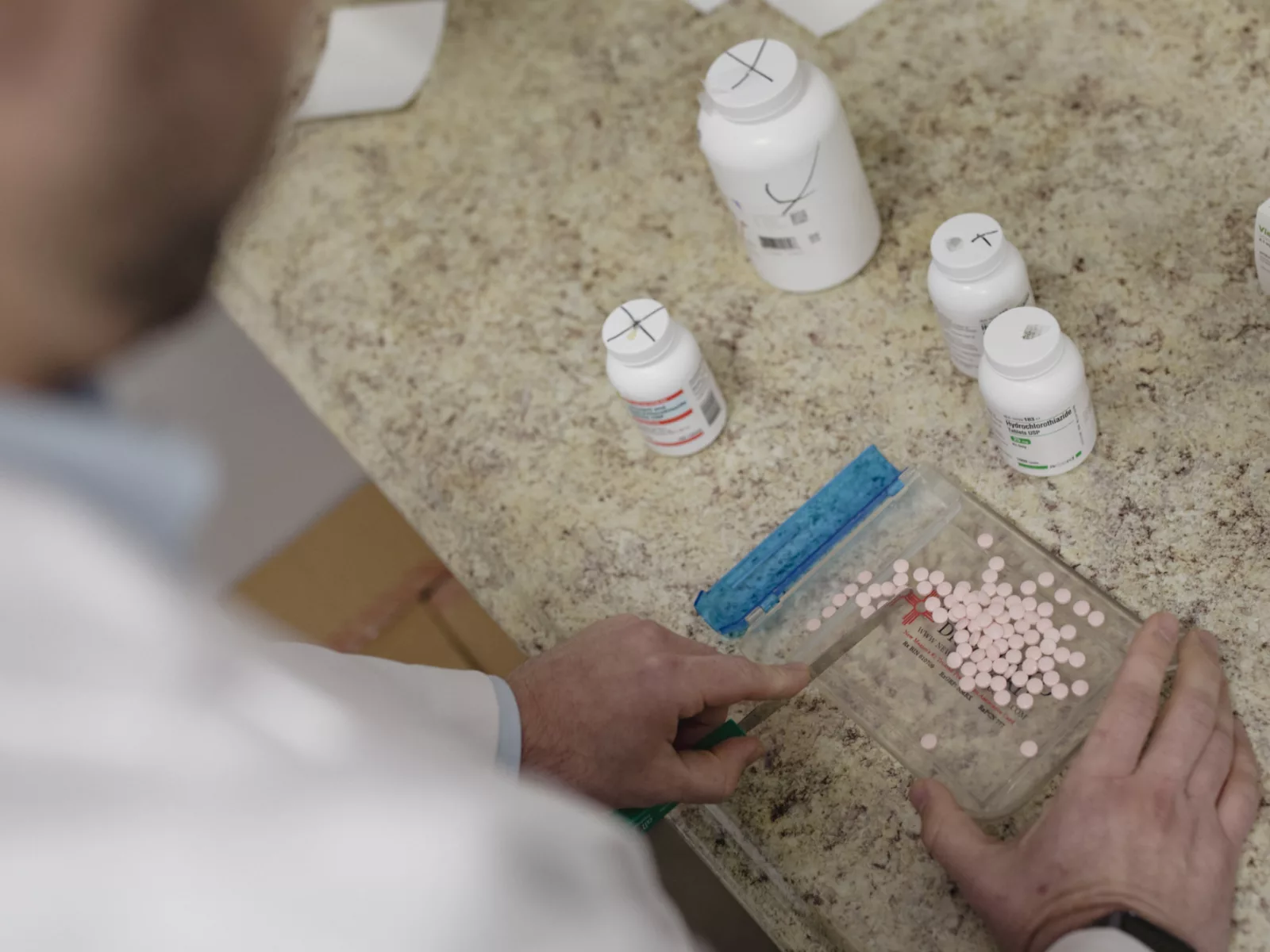 A healthcare professional in a lab coat is counting pink tablets on a tray, surrounded by various bottles of medication on a countertop.