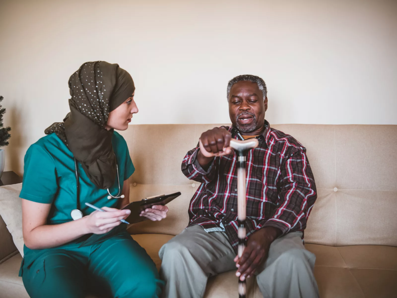 Woman in scrubs sits on couch with a patient holding a cane.