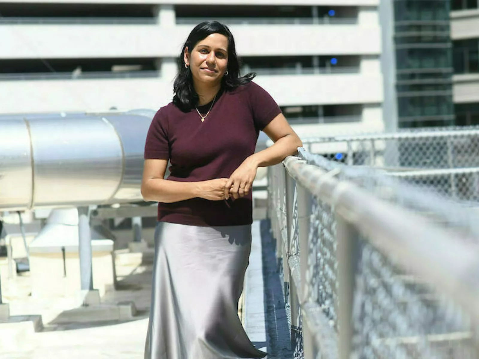 Woman stands on rooftop of building leaning on chain link fence.