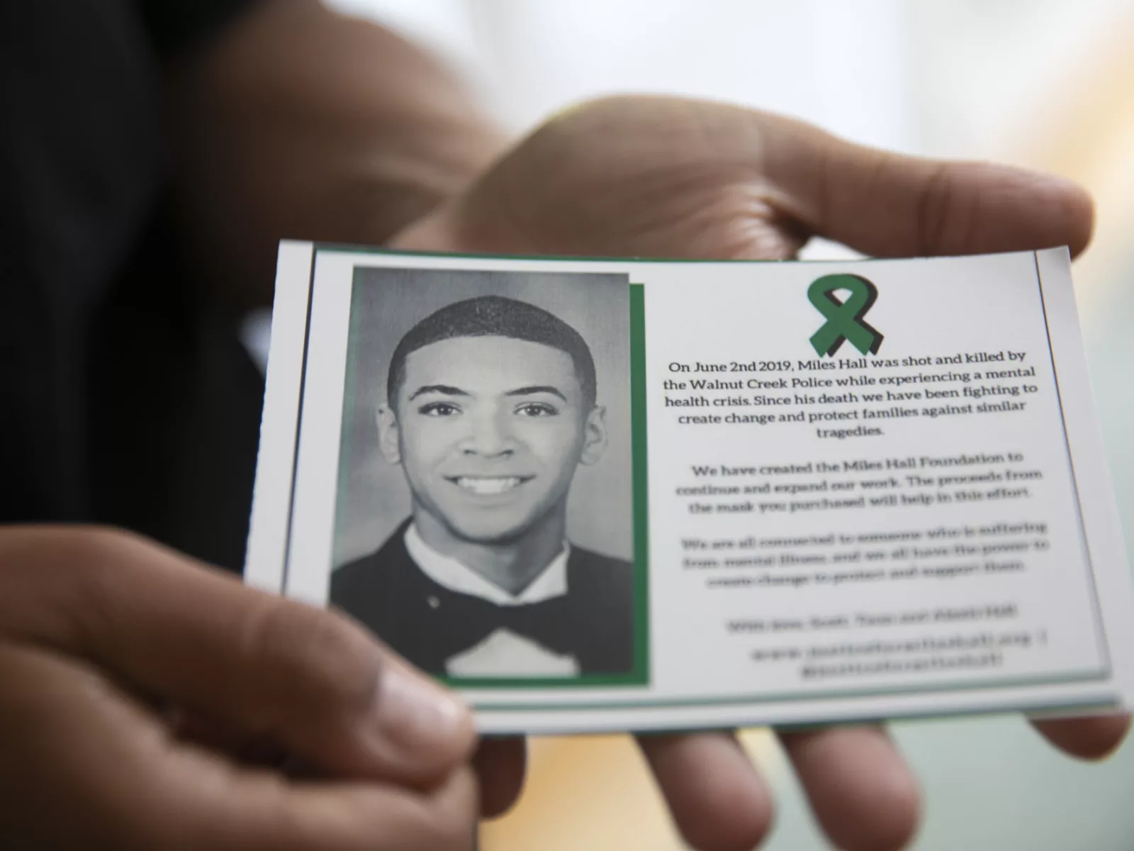 A hand holds a card honoring Miles Hall, featuring his photo, a green ribbon, and text about addressing mental health issues after his tragedy.