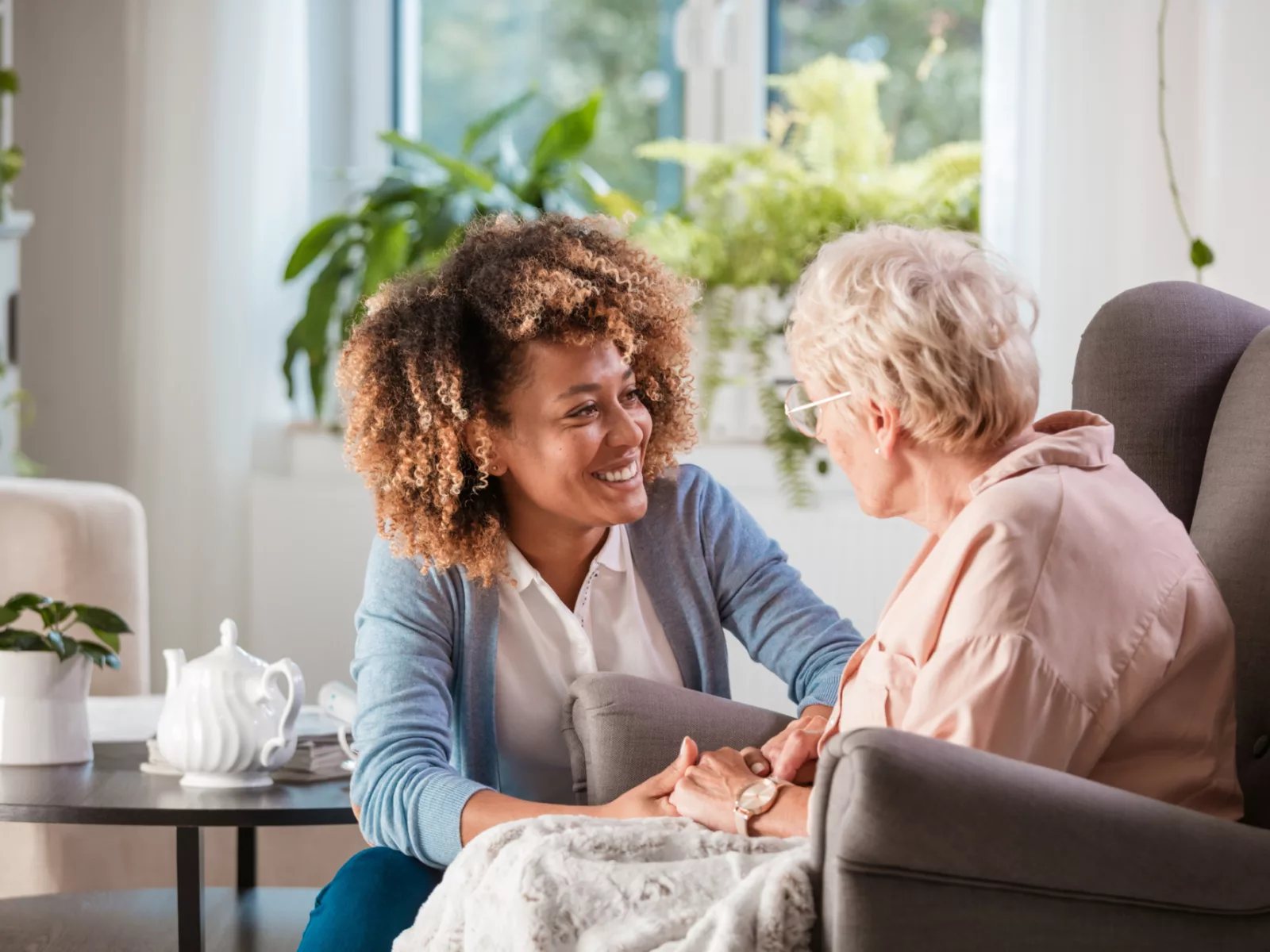 A young woman with curly hair smiles warmly while holding the hands of an elderly woman in a cozy, plant-filled room.