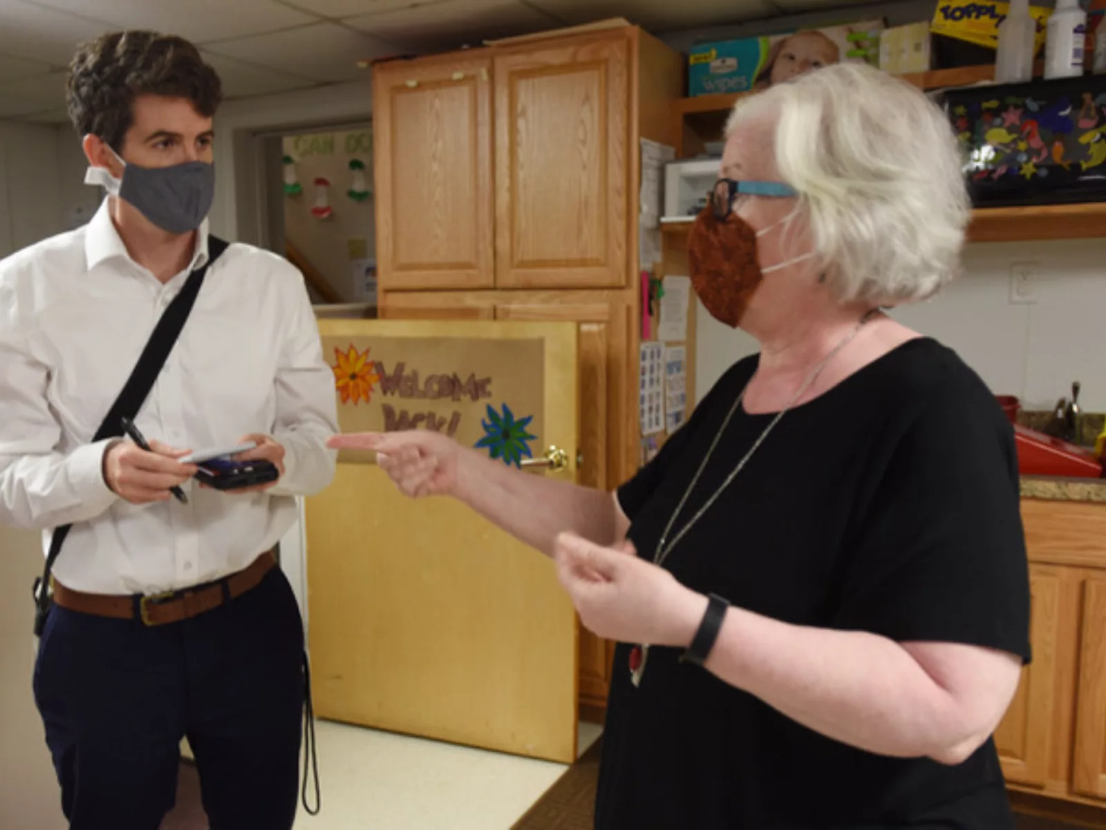 A reporter with a notebook and a face mask stands in the kitchen takes notes while interviewing a woman who gestures with her hands.