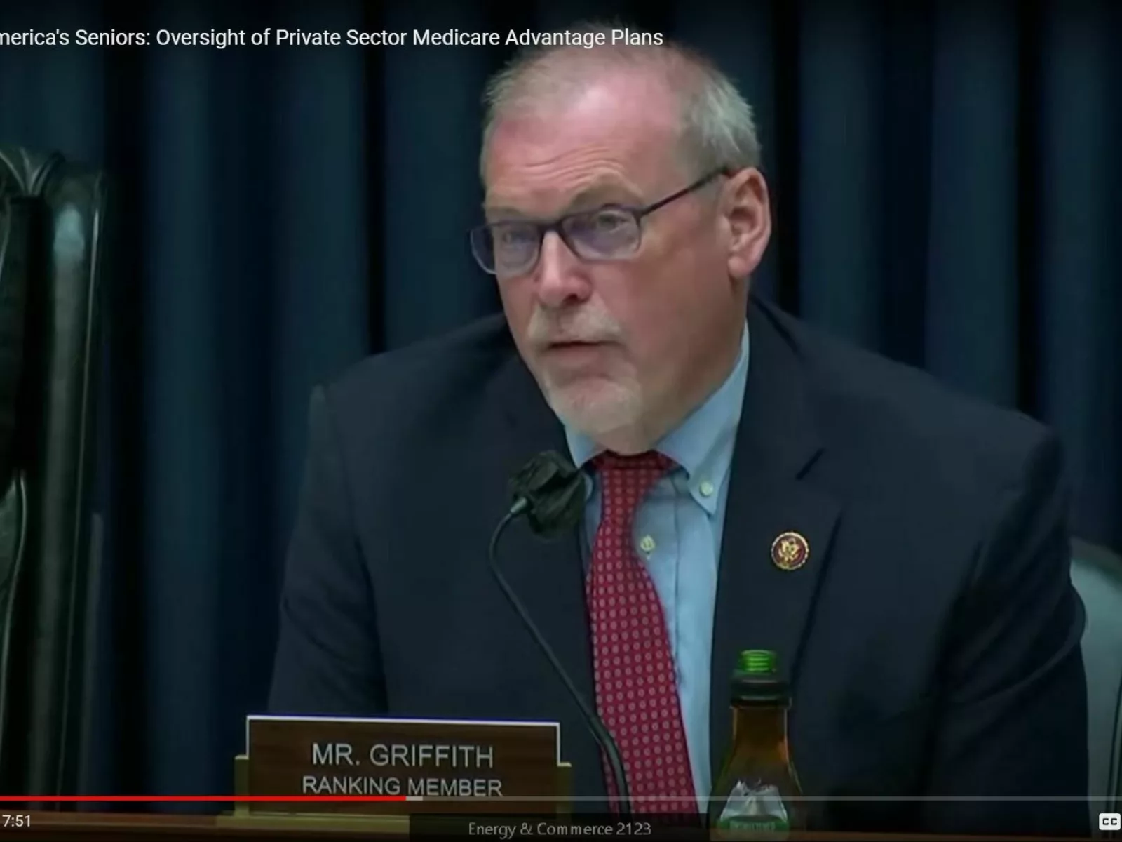 A man in a suit with glasses speaks at a hearing, sitting at a table with a microphone and a green beverage bottle nearby.