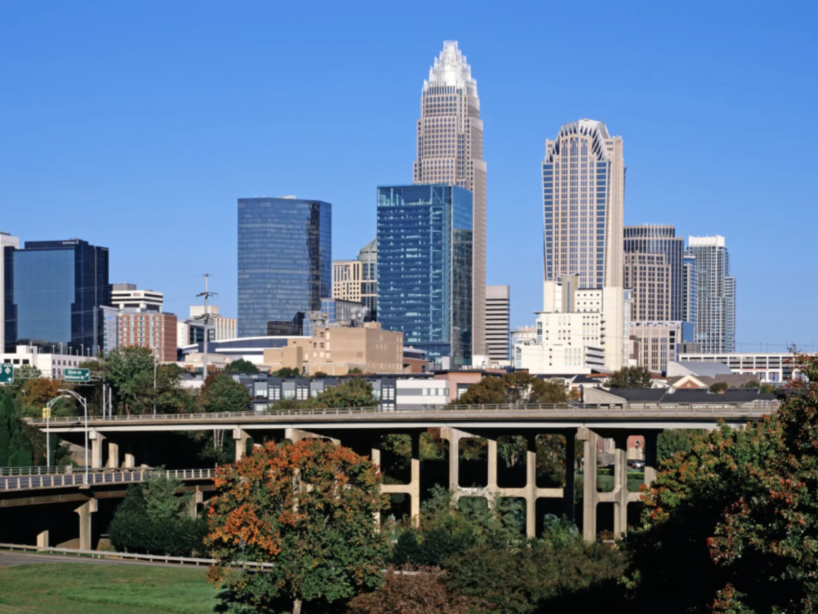 Skyline of Charlotte, North Carolina shows skyscrapers, trees and highways against a bright blue sky.