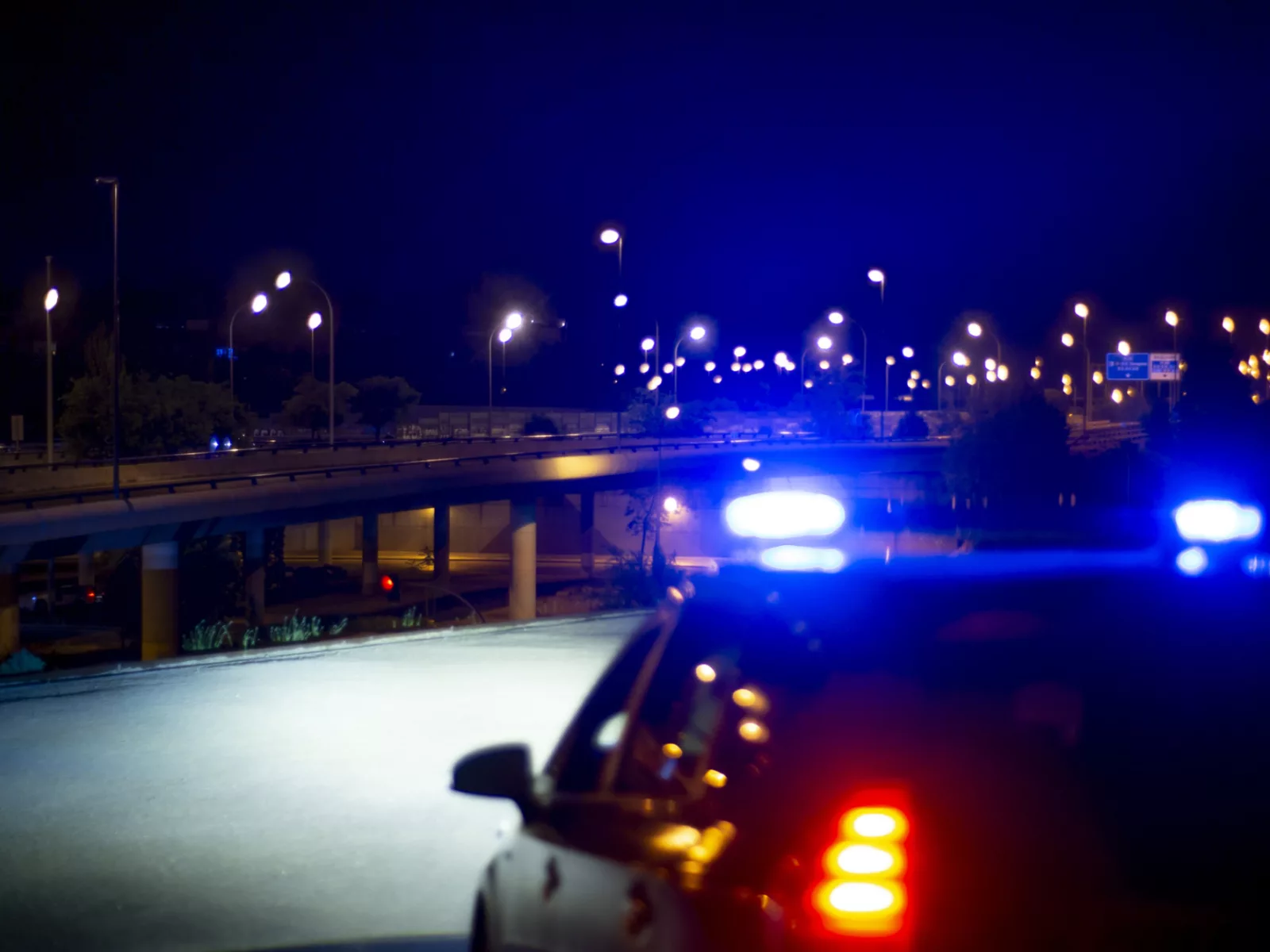 Emergency lights illuminate a dark urban highway at night, creating a vibrant contrast against the calm surroundings and distant streetlights.