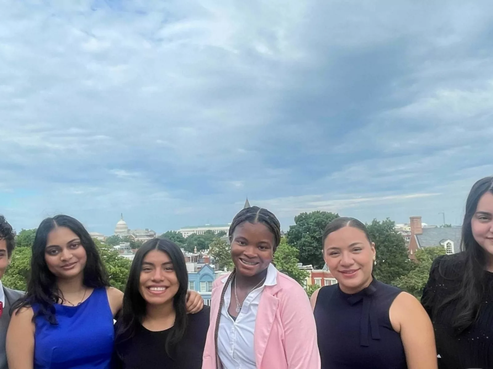 A diverse group of six young adults stands together outdoors, smiling, with the U.S. Capitol visible in the background under a cloudy sky.
