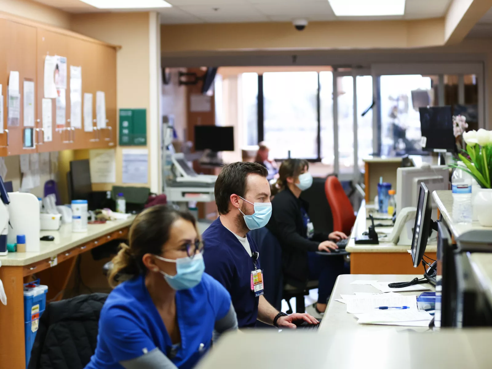 Healthcare professionals in scrubs and masks work at computers in a busy hospital environment, with documents and plants on the counter.