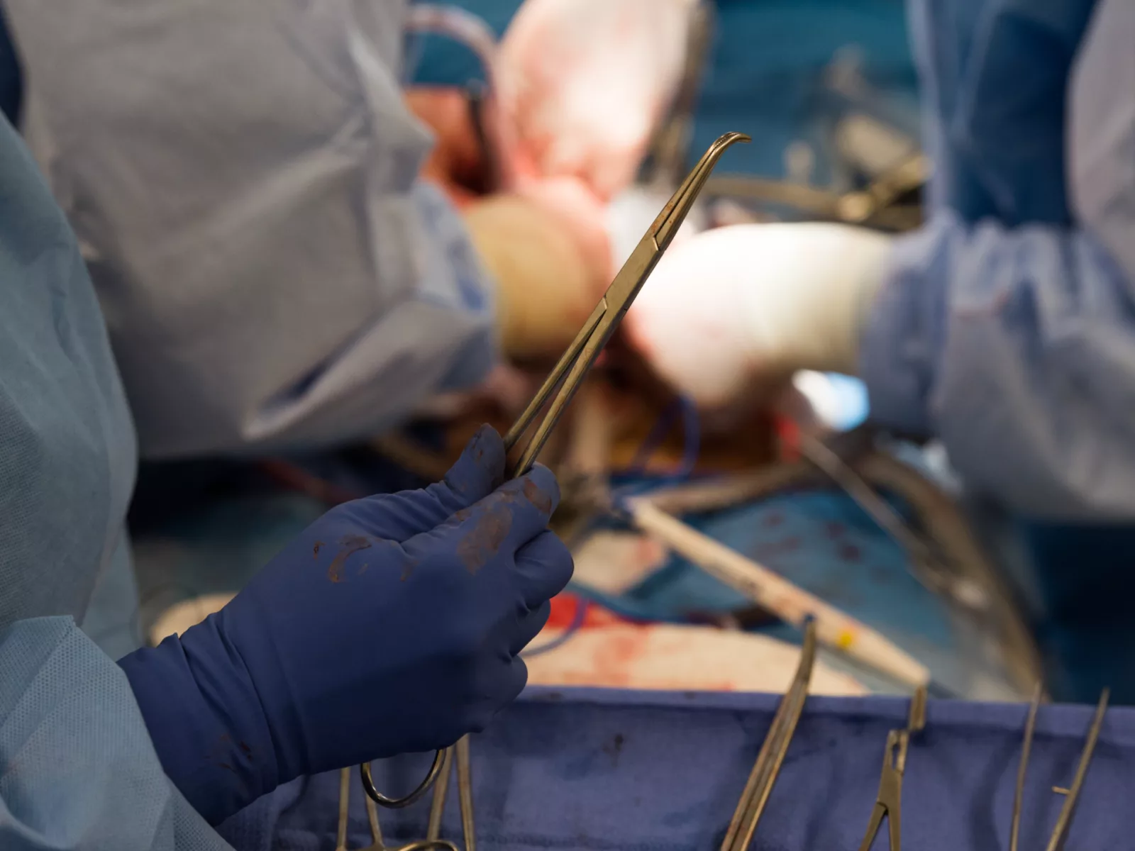 Hands in blue surgical gloves hold a surgical instrument, surrounded by various tools and a blurred surgical field in the background.