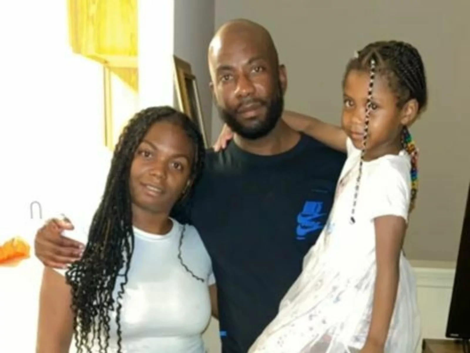 A woman and a man stand together, smiling, with two young girls; one girl is in the man's arms and the other is playfully posing.