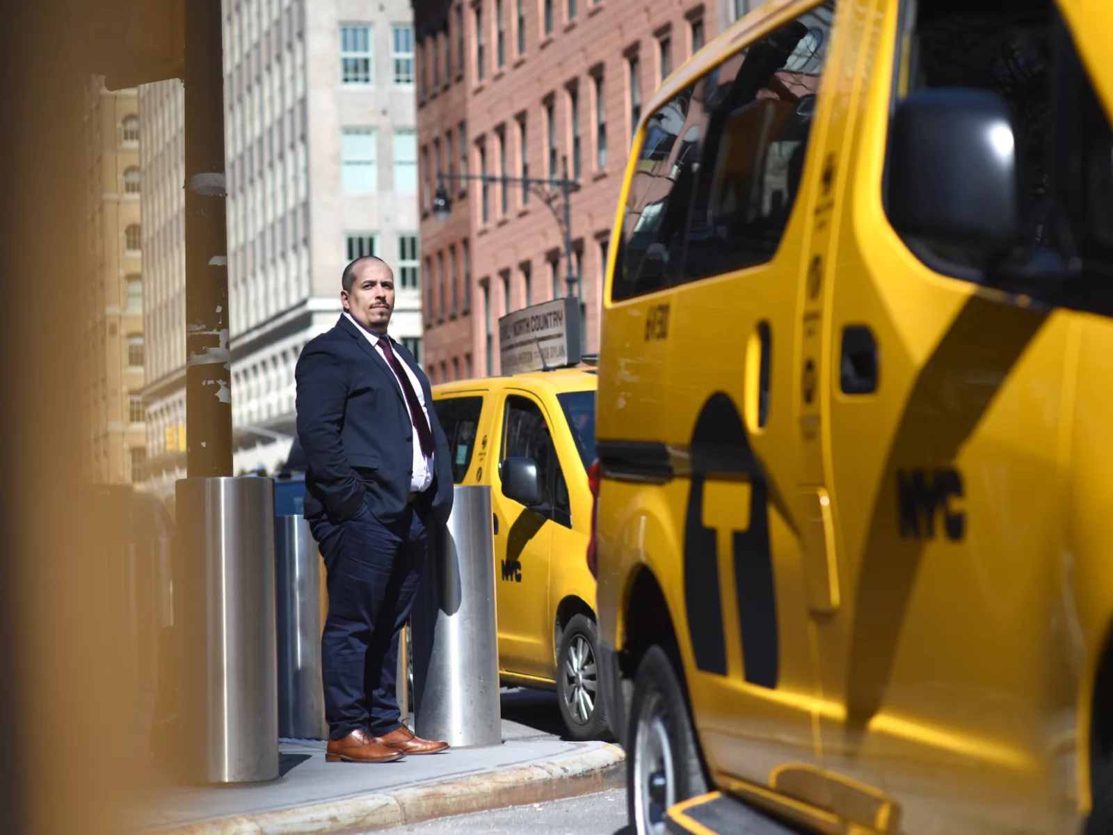 A man in a suit stands beside a yellow taxi, with city buildings in the background. Sunlight brightens the urban scene.