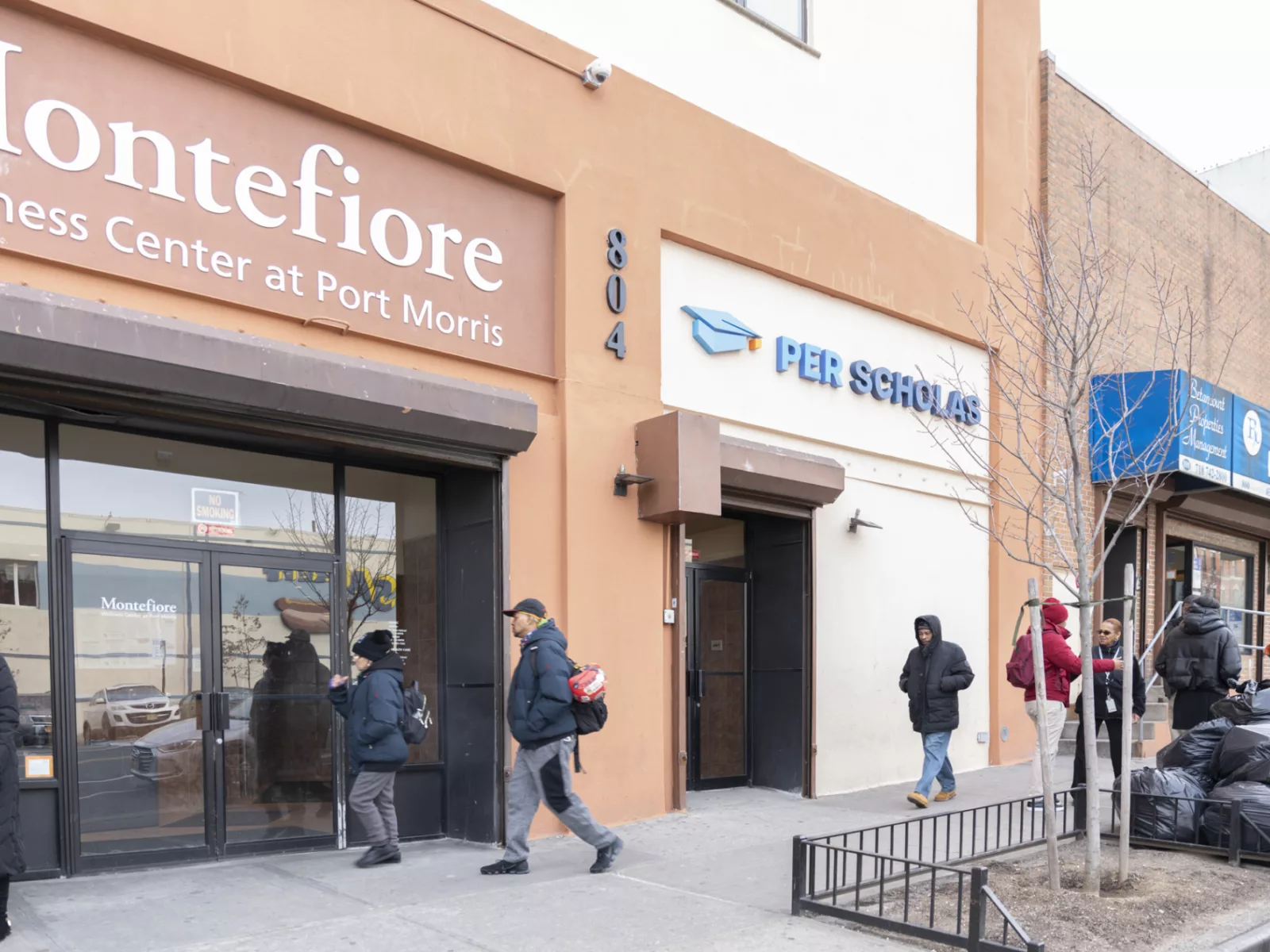 A bustling city street featuring Montefiore Wellness Center and Per Scholas, with pedestrians walking past and a pharmacy nearby.