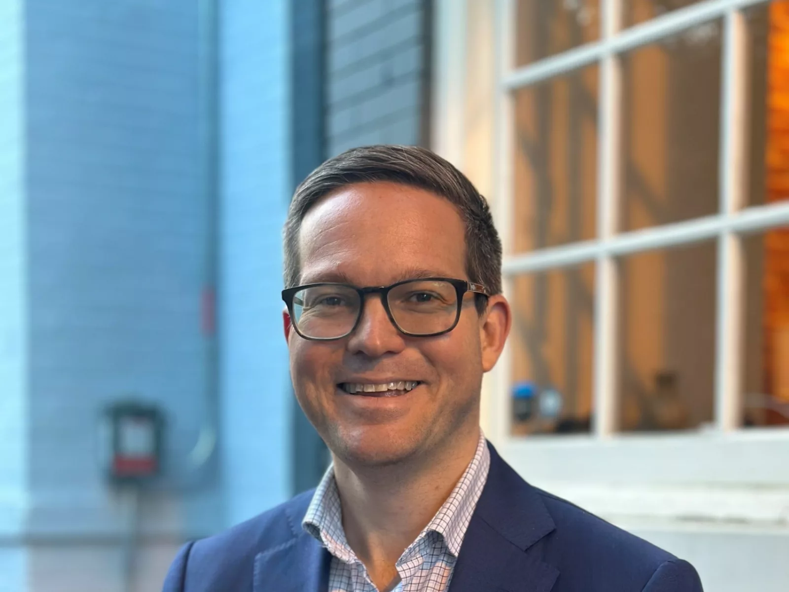 Smiling man in a tailored navy suit stands outside, with a light blue textured wall and a softly lit window in the background.