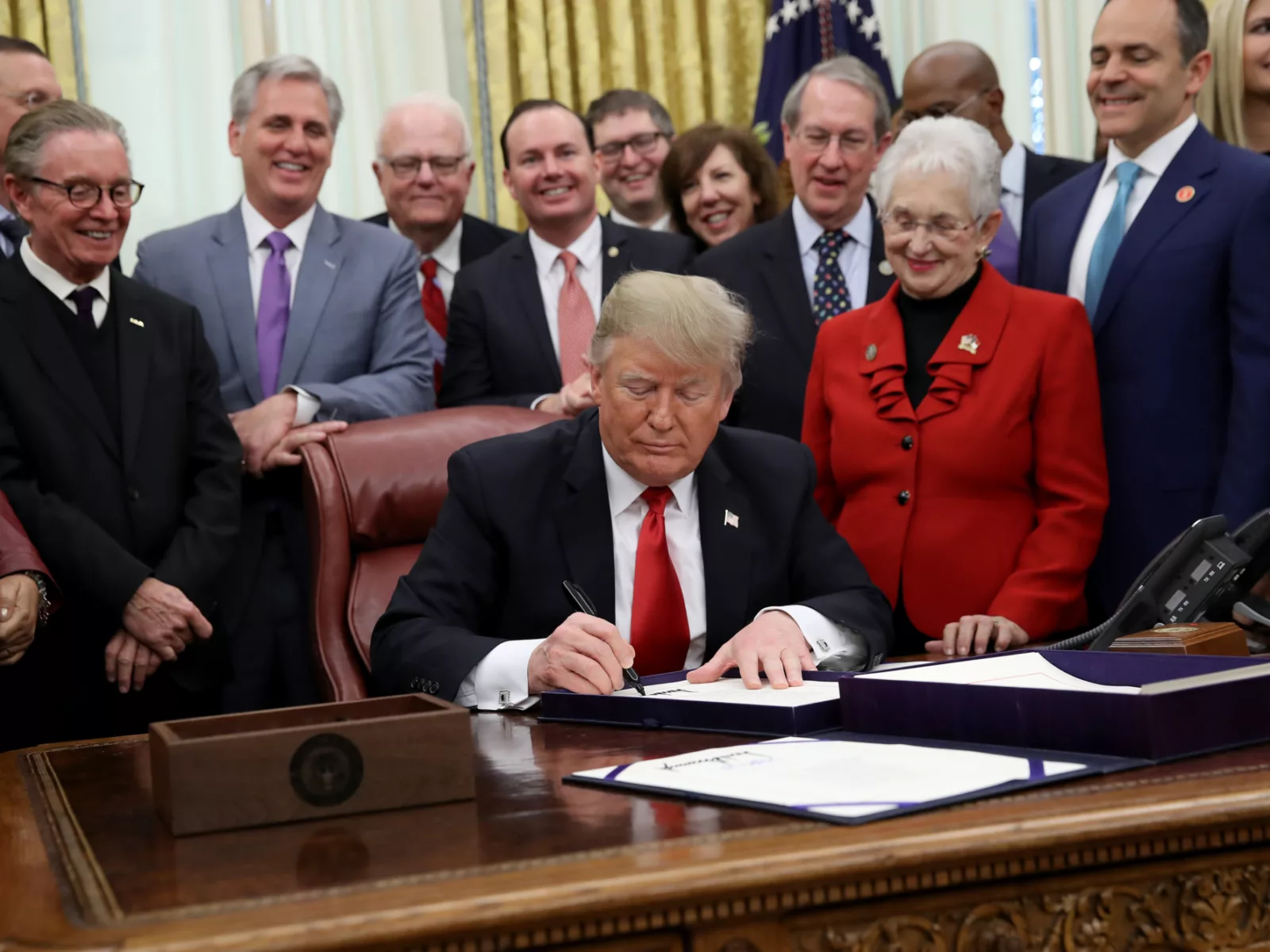 Donald Trump signs a document at a desk, surrounded by smiling officials in suits, capturing a moment of legislative celebration.