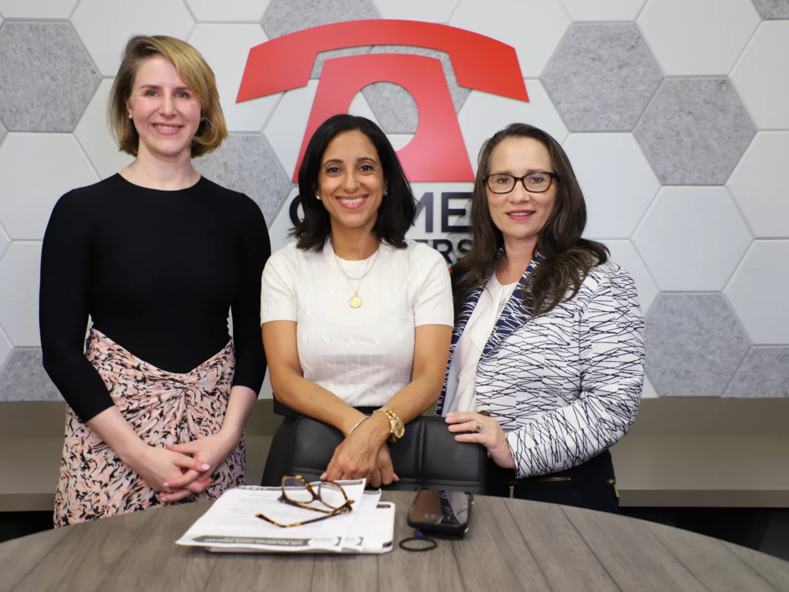 Three women stand together, smiling, in a modern office setting with a hexagonal wall design. Papers and glasses are on the table.