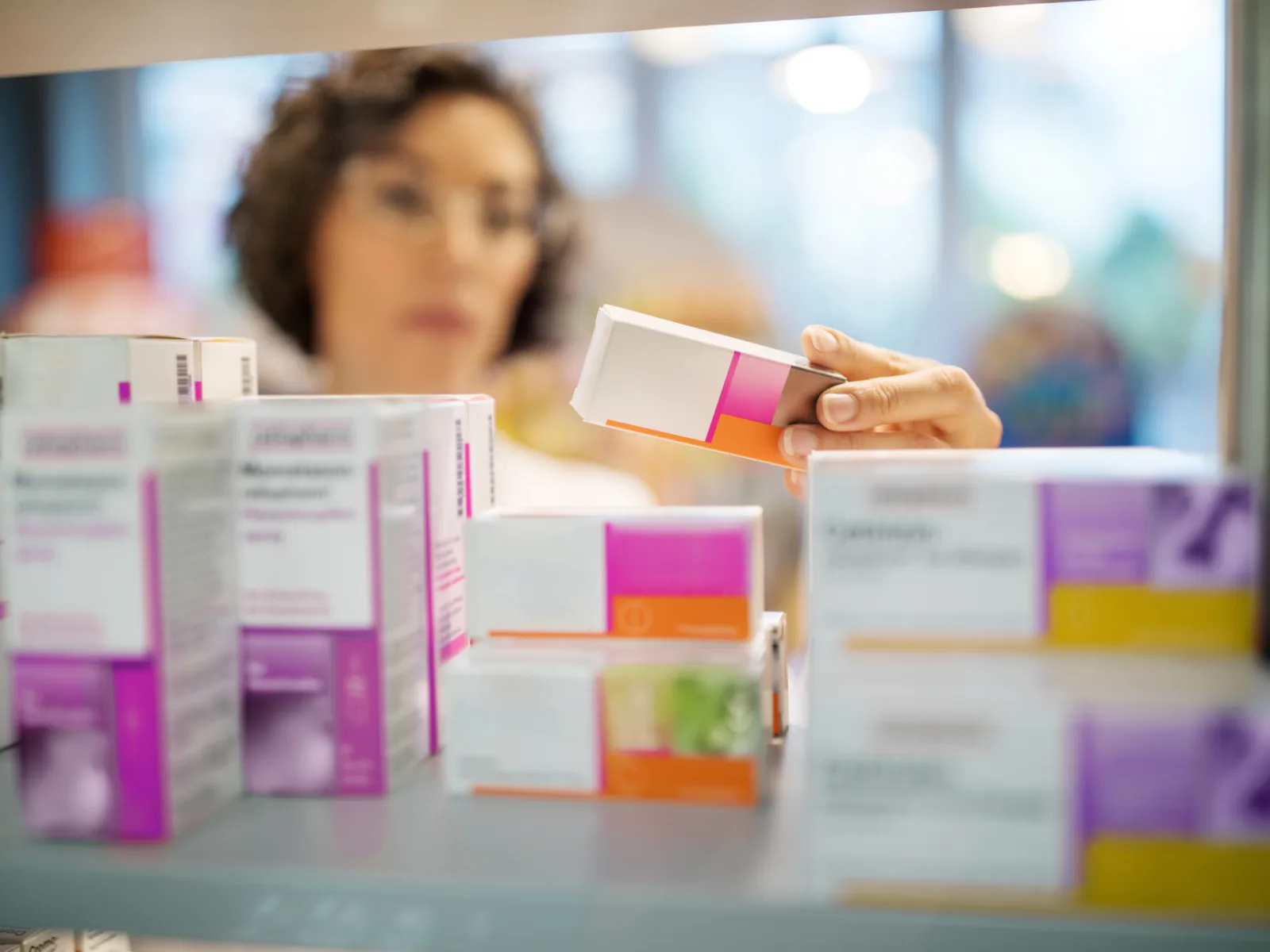 A pharmacist examines a medication box with pink accents while shelves filled with various other pharmaceutical products are visible.