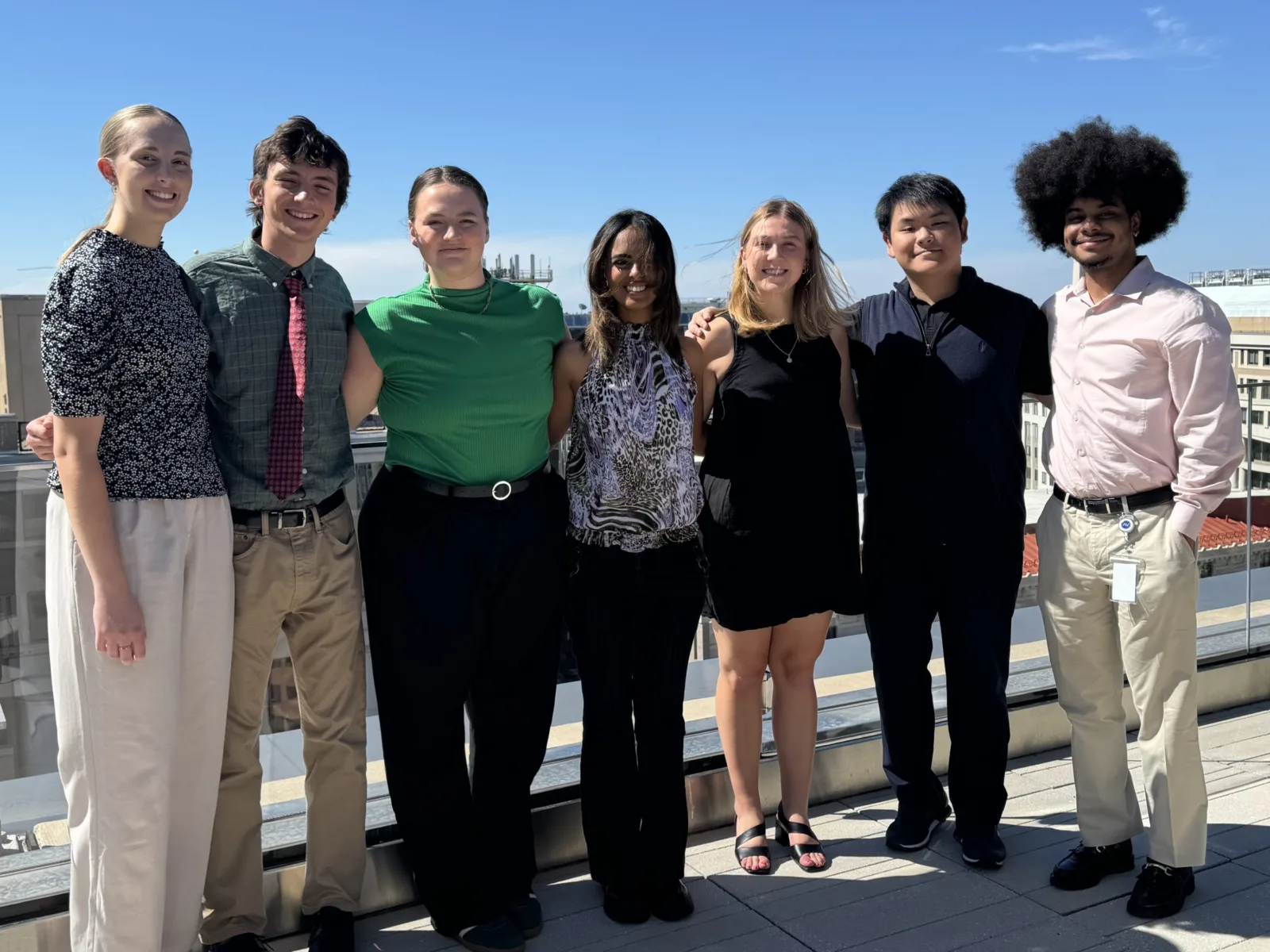 A diverse group of six young adults stands together outdoors under a clear blue sky, smiling and dressed in stylish outfits.