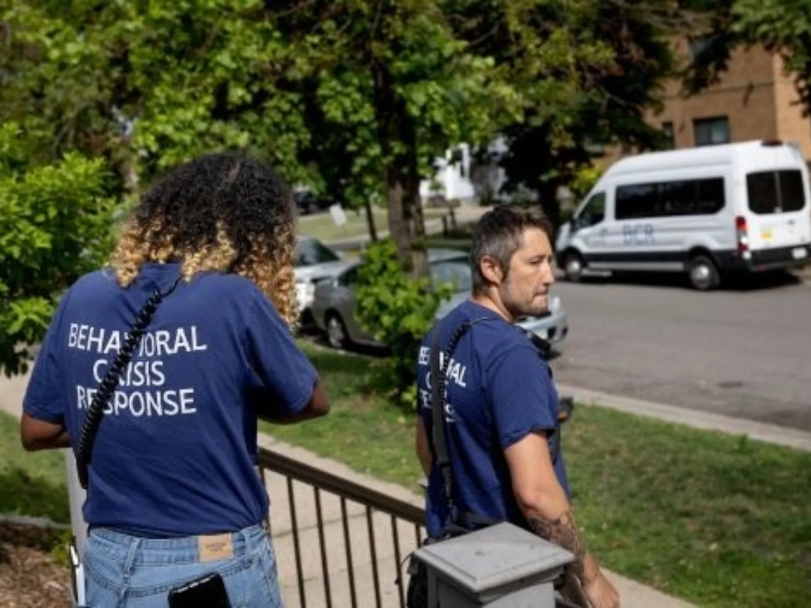 Two members of a behavioral crisis response team, wearing navy shirts, walk down steps towards a residential street with trees.