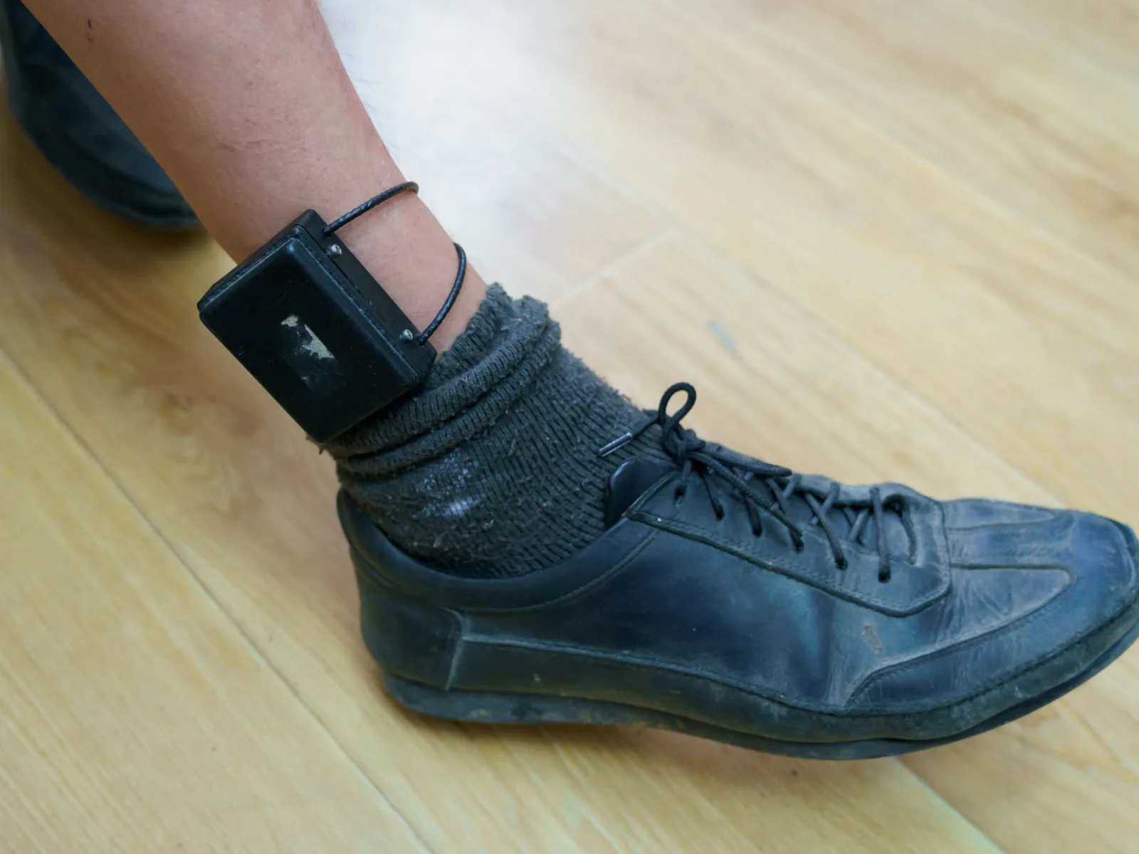 A black electronic monitoring device is attached to a person's ankle, alongside a black shoe and dark sock on a wooden floor.