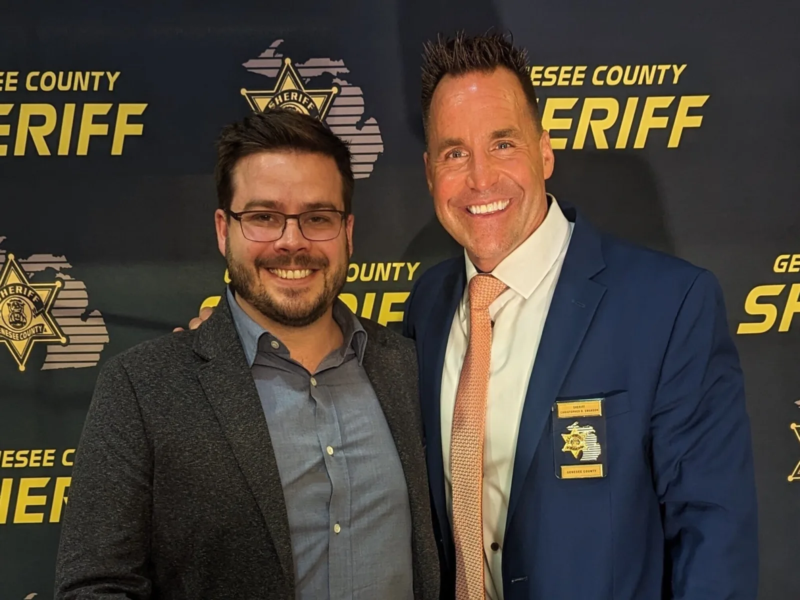 Two men pose together, one in a gray blazer and glasses, the other in a blue suit with an orange tie, smiling against a sheriff's backdrop.