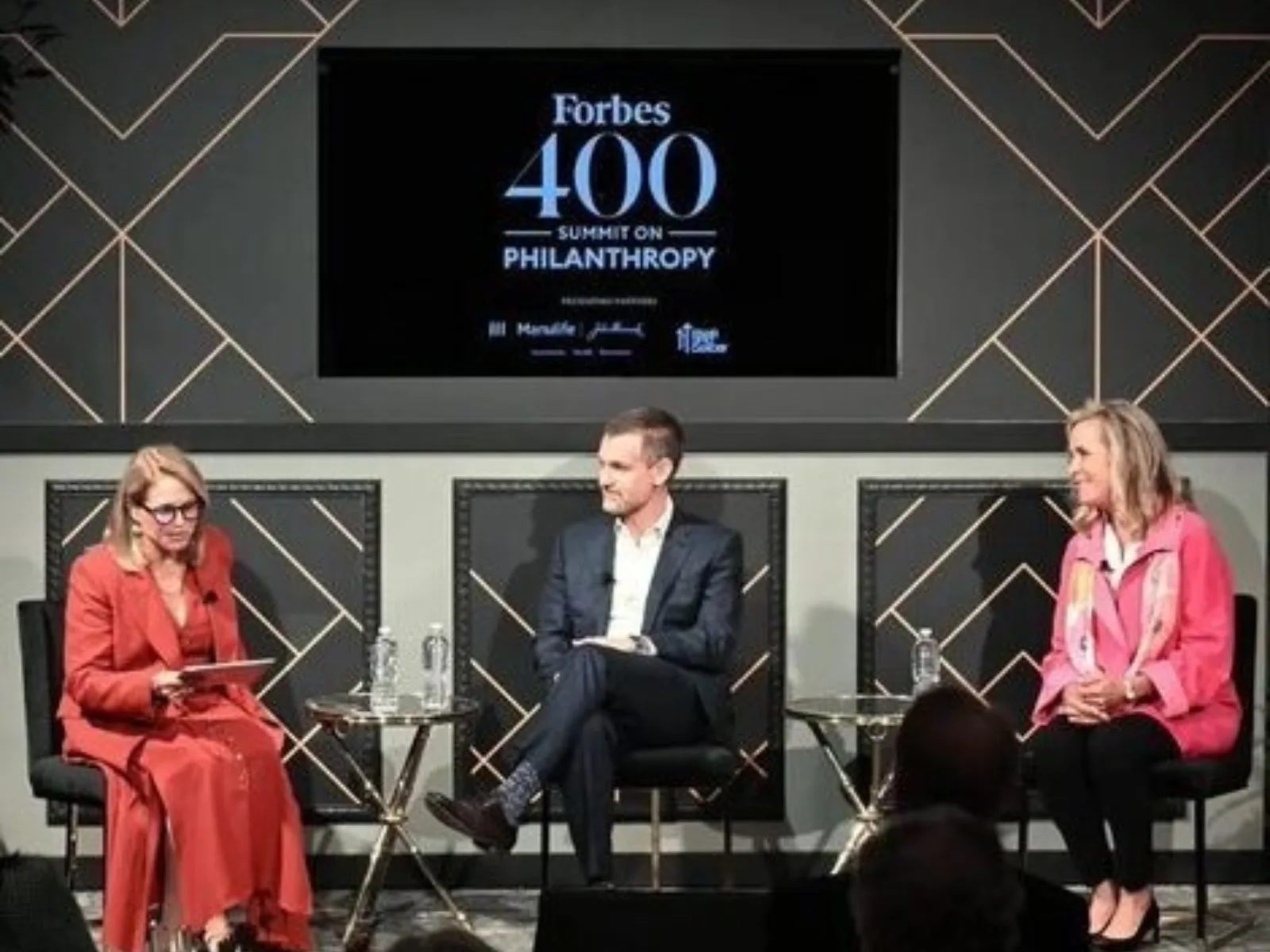 A panel discussion featuring three speakers: a woman in a red dress, a man in a suit, and a woman in a pink scarf, discussing philanthropy.