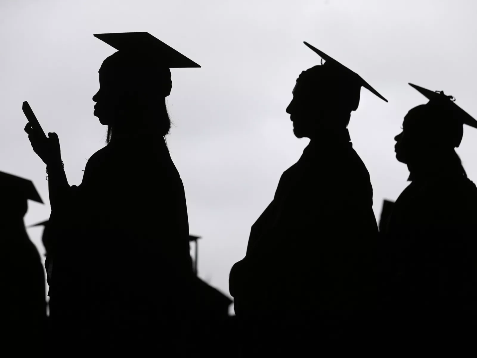 Silhouettes of graduates wearing caps stand in profile, one checking a phone, against a cloudy sky, capturing a moment of anticipation.