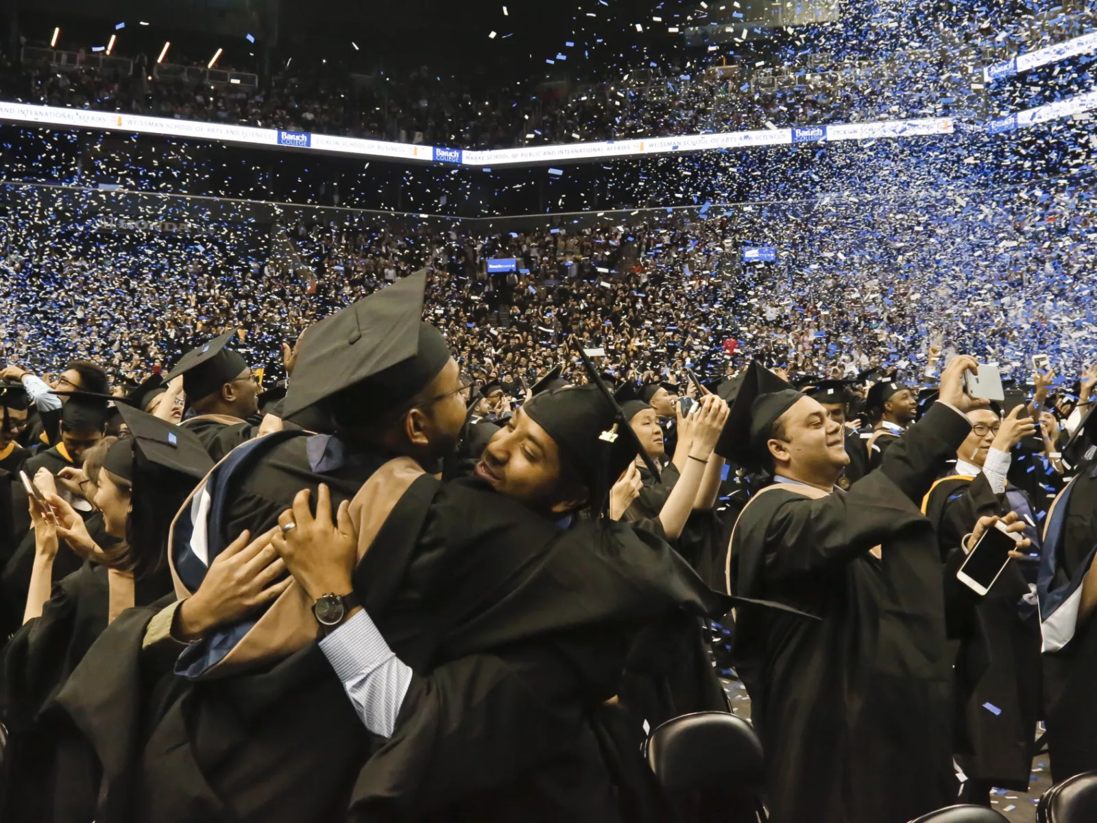 Graduates in caps and gowns celebrate at a ceremony, sharing hugs amid a shower of blue confetti and capturing moments on their phones.