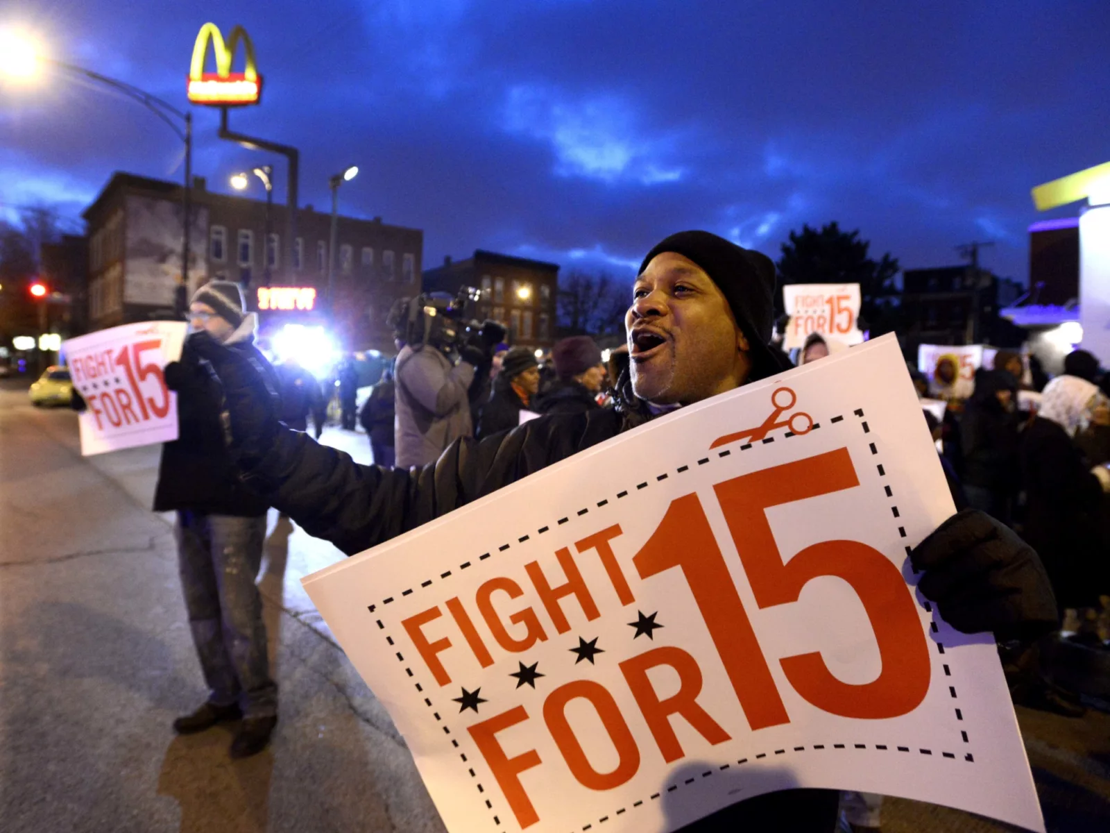 A crowd rallies at dusk, holding signs that read "Fight for 15," advocating for higher wages, with a McDonald's sign in the background.