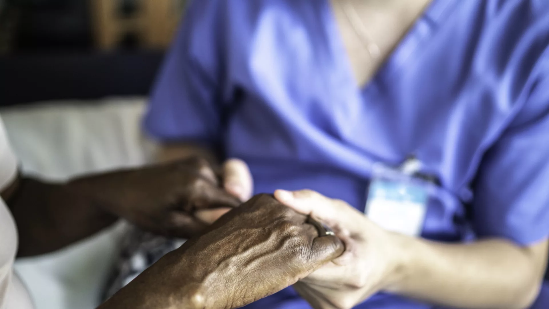 Two hands are clasped together, one belonging to a healthcare professional in blue scrubs, the other an older individual with dark skin.