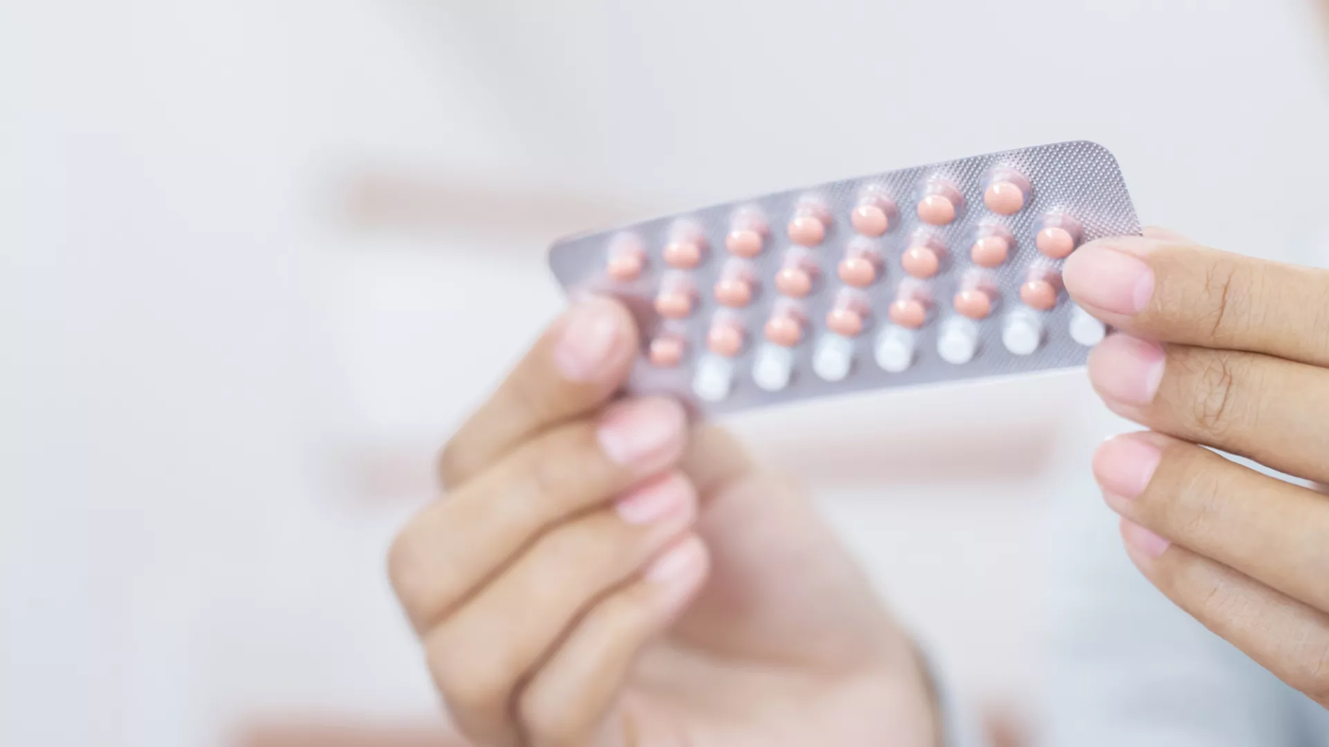 A hand holds a blister pack of birth control pills, featuring several pink and white tablets, against a soft, blurred background.