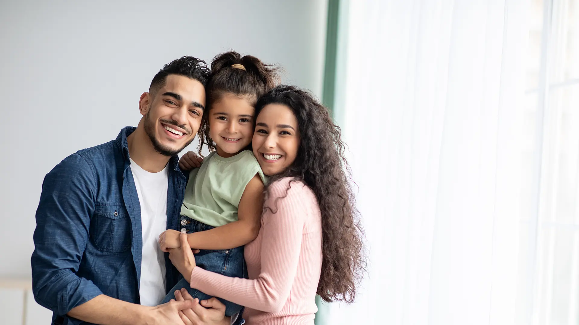 A smiling couple holds their young daughter between them, radiating happiness in a bright room filled with natural light.