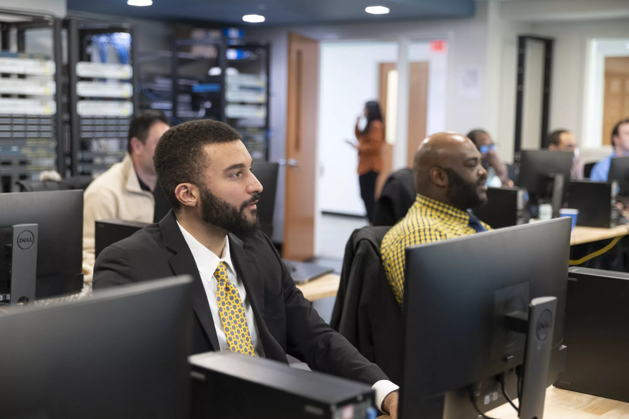 Two men are seen sitting at a long table with computers in front of them during a skills training class.
