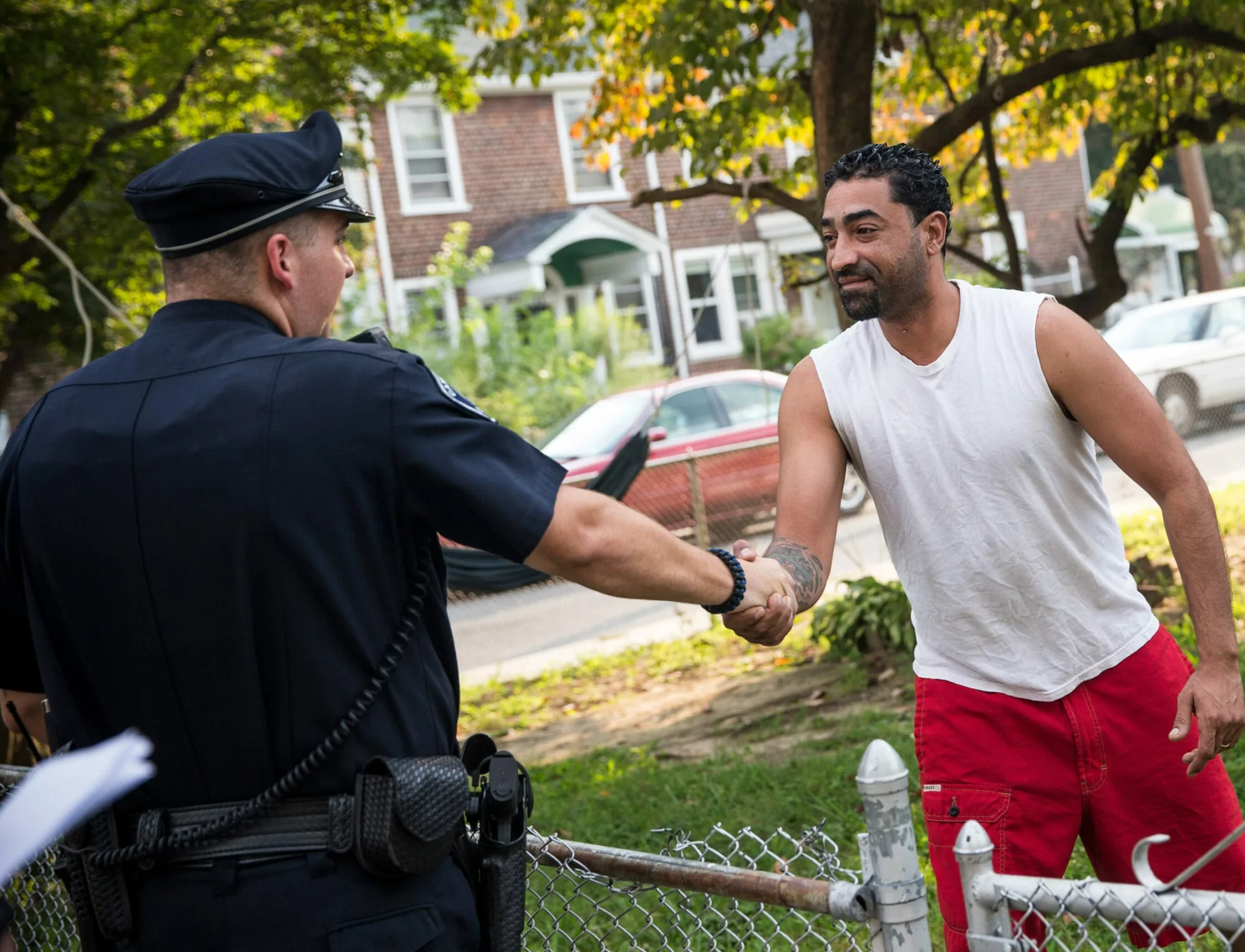 A police officer shakes hands with a male resident over a chain link fence in a residential neighborhood.