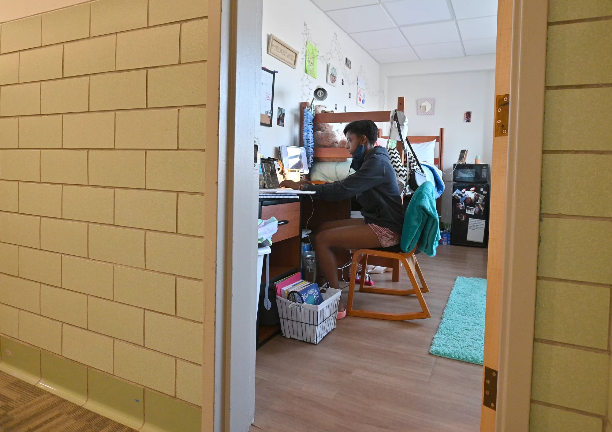 Student is seen sitting at her desk working on a laptop in her dorm room through the hallway door.