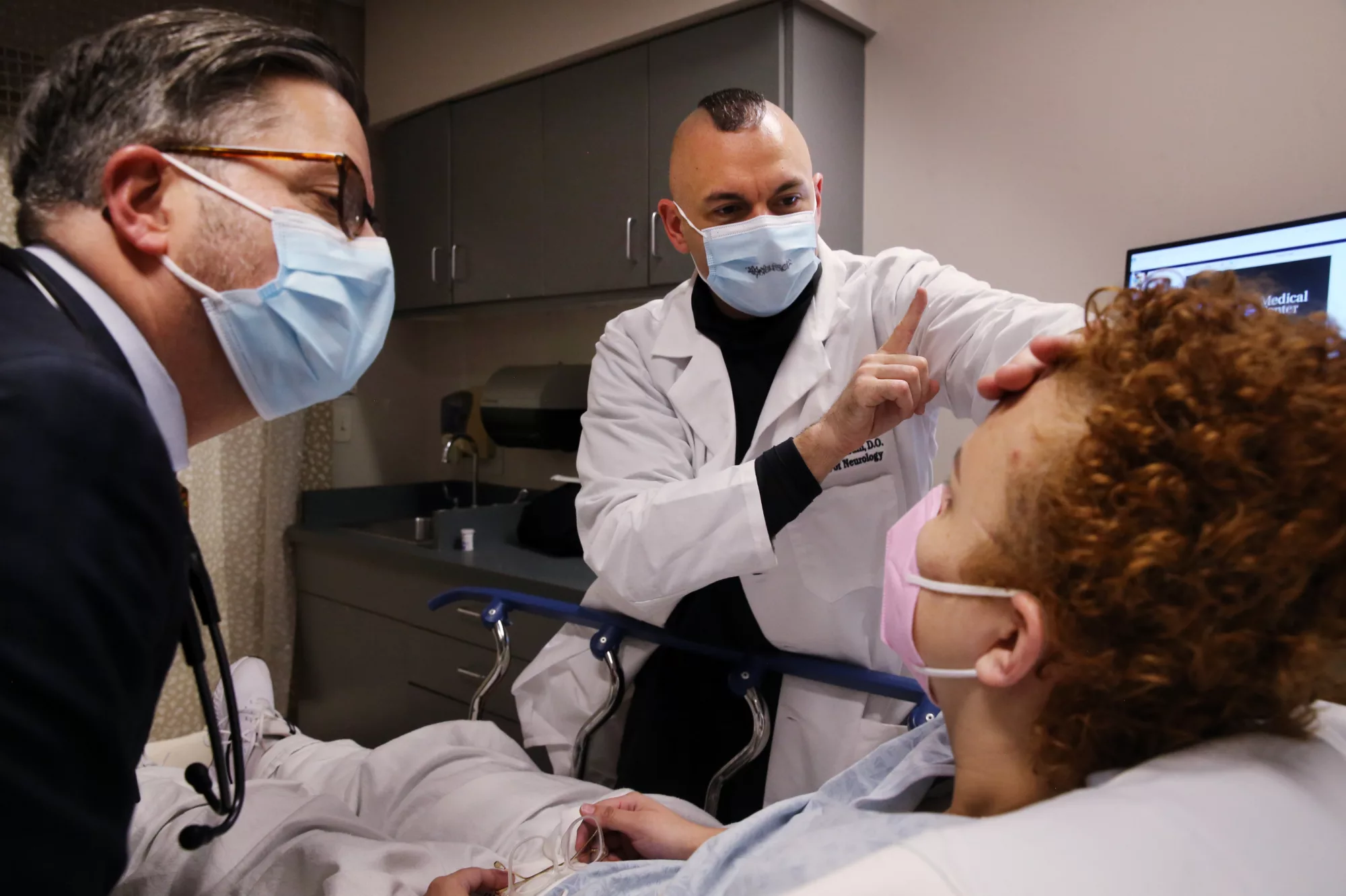 Two doctors wearing face masks lean over either side of a patient lying down in an ER bed, with one doctor holding up an index finger to the patient at eye level.