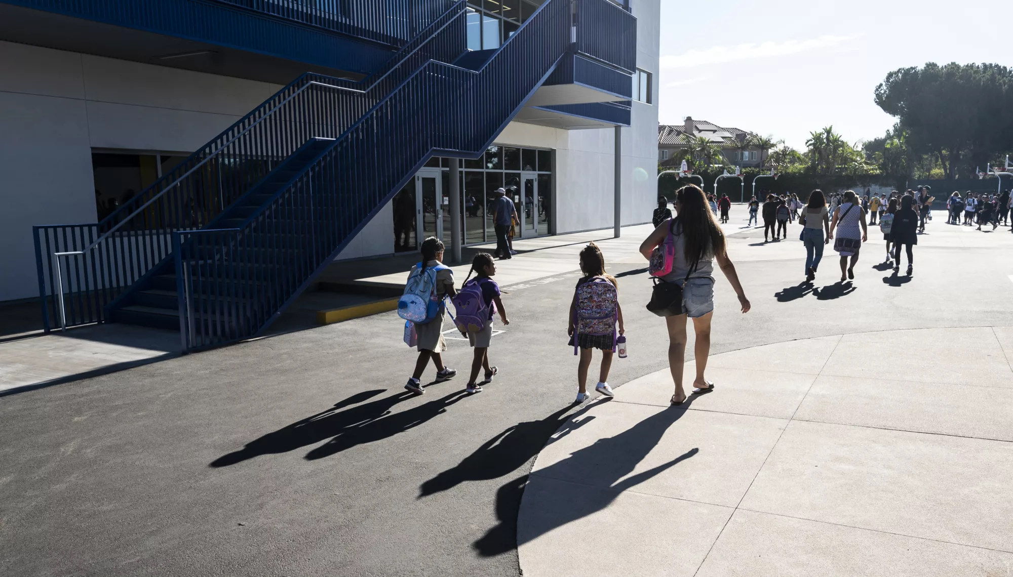 Students wearing backpacks walk with families next to a school building on a bright morning casting shadows against the concrete.