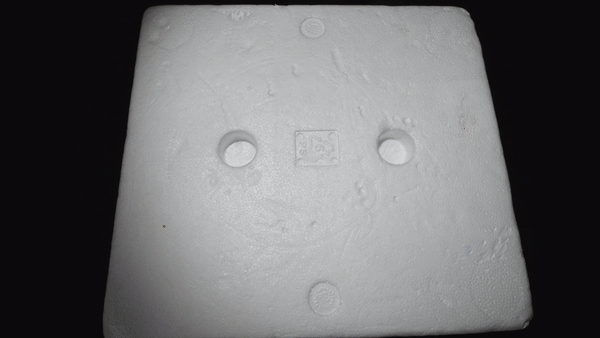 A textured, white foam sheet features two circular indentations and a small square logo in the center, set against a black background.